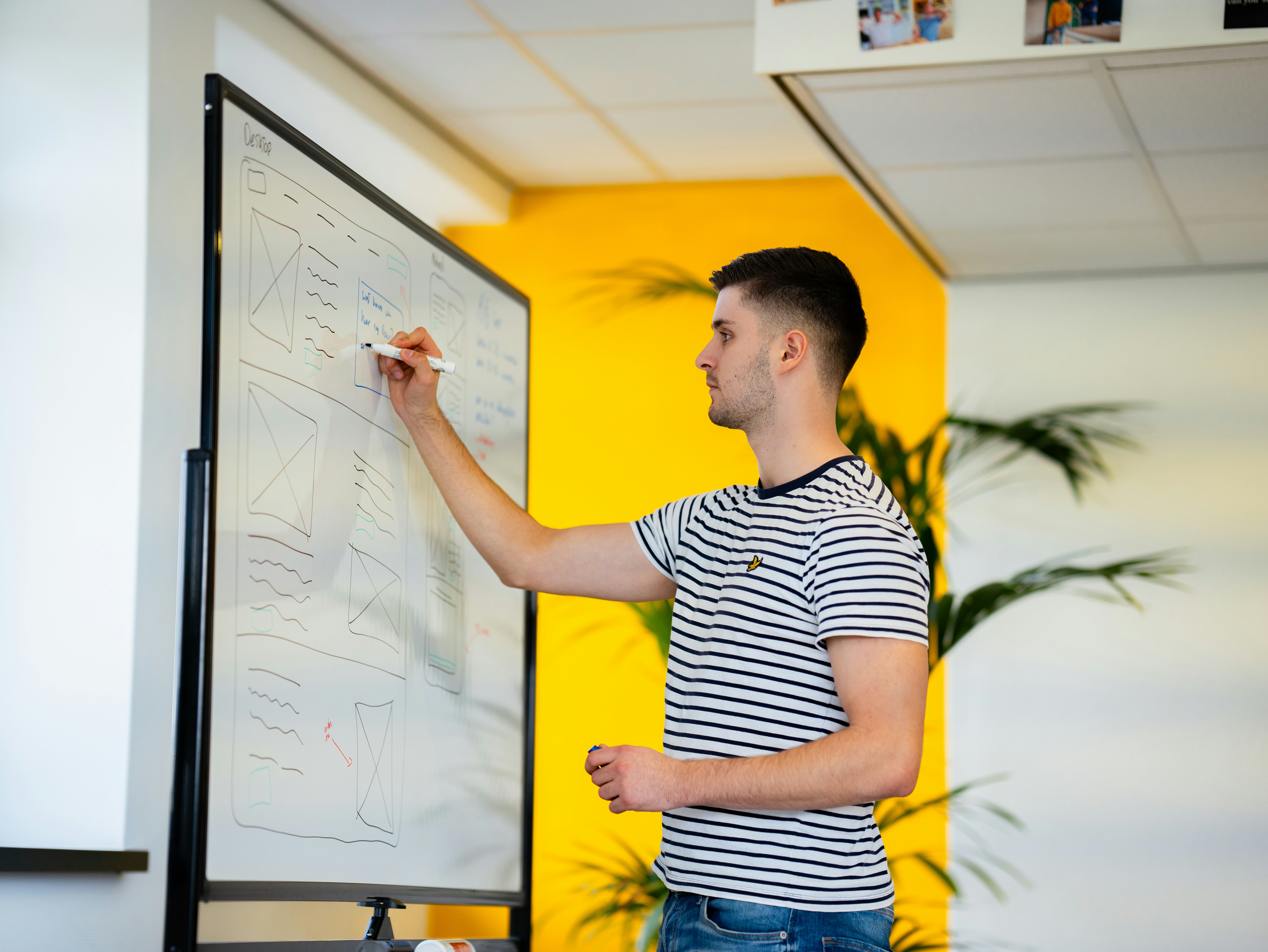 Young man in a striped shirt writing on a whiteboard in a bright office space.