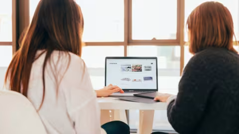 Two women sitting at a table, working on a laptop in a bright room with large windows.